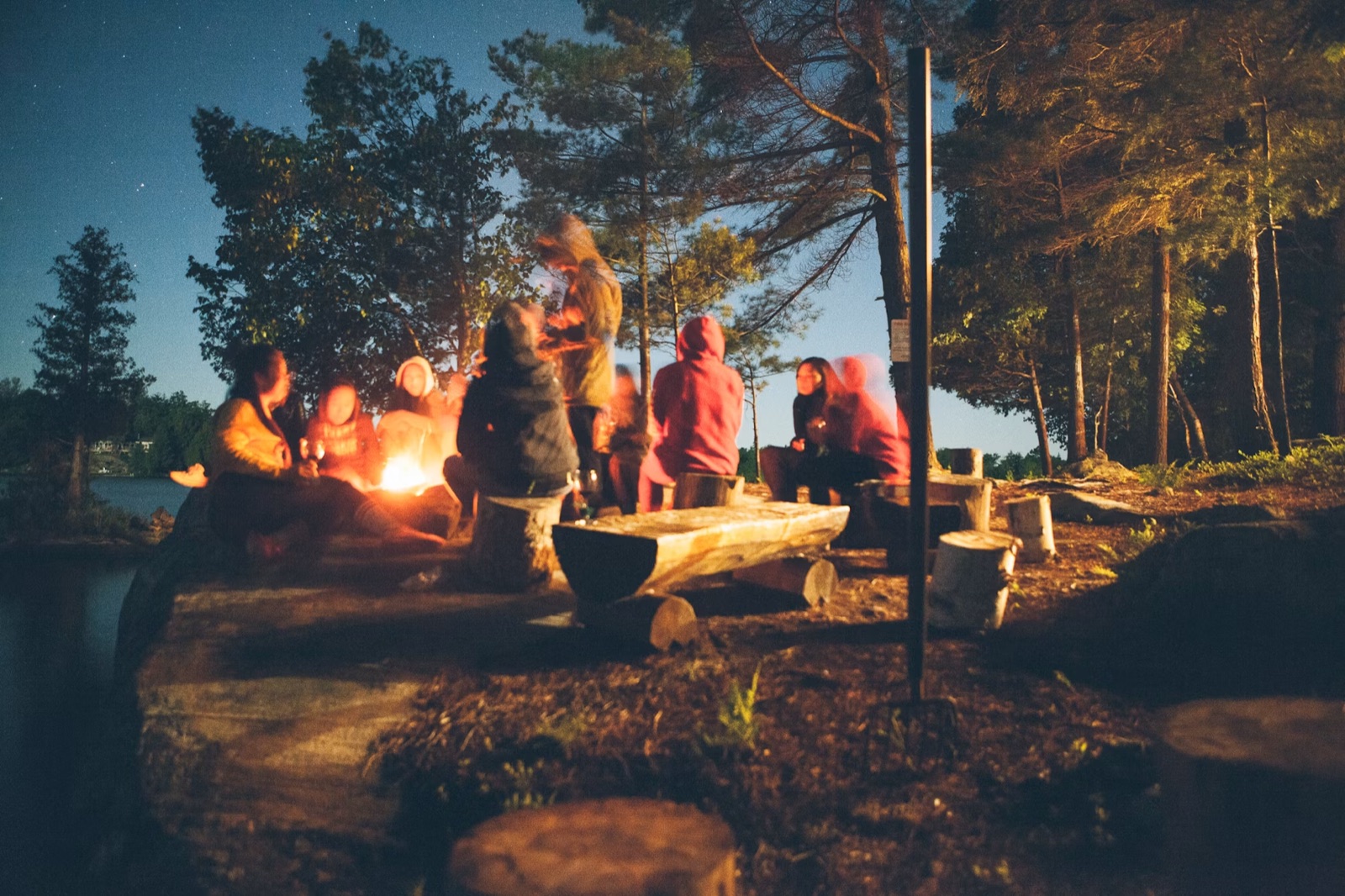 Friends gathered around a glowing campfire at dusk by the water.